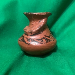 Puede incluir: Un pequeño jarrón de terracota pintado a mano, con un cuerpo bulboso y un borde acampanado. El jarrón presenta un color base marrón oscuro con diseños abstractos negros alrededor del cuerpo. Un asa decorativa se curva alrededor del cuello.