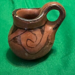 May include: A small, handcrafted ceramic pitcher with a rounded body and a loop handle. The pitcher is primarily brown with dark brown, abstract designs. The rim and handle are outlined in a darker shade. The pitcher sits on a green surface.