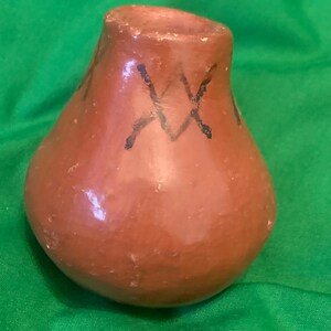 May include: A small, brown, handmade ceramic vase with a rounded body and a slightly flared opening. The vase is decorated with black, X-shaped markings. The background is a solid green.