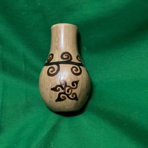 May include: A small, beige ceramic vase with a narrow neck and rounded body. The vase is decorated with black swirl and floral designs. The vase is sitting on a green surface.