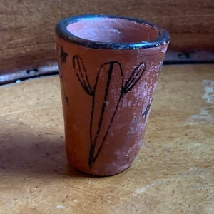 May include: Small, hand-painted terracotta cup with a black rim and a design of black lines and shapes. The cup has a rustic, aged appearance, with visible texture and variations in the reddish-brown color.