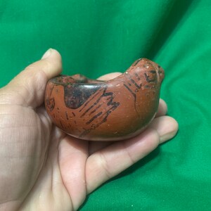 May include: A small, hand-held, terracotta-colored ceramic bowl with a bird-like shape. The bowl features black painted details, including what appears to be a bird design on the side. The bowl is held in a person's hand against a green background.