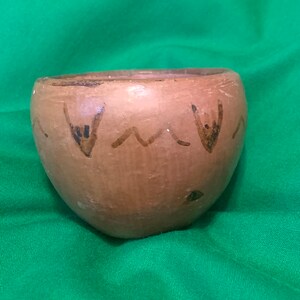 May include: A small, handmade, brown ceramic bowl with a rounded shape. The bowl is decorated with dark brown and black painted designs, including angular shapes and wavy lines. The bowl sits on a green fabric surface.