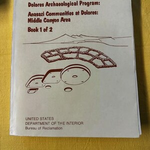May include: A book cover with the title "Dolores Archaeological Program: Anasazi Communities at Dolores: Middle Canyon Area Book 1 of 2." The cover features a brown illustration of a landscape and structures. The bottom of the cover reads "UNITED STATES DEPARTMENT OF THE INTERIOR Bureau of Reclamation."