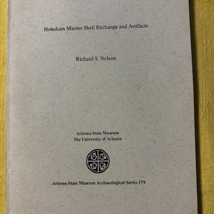 May include: A book titled "Hohokam Marine Shell Exchange and Artifacts" by Richard S. Nelson. The cover features text and a circular emblem, with the Arizona State Museum and The University of Arizona logos. The book is part of the Arizona State Museum Archaeological Series 179.