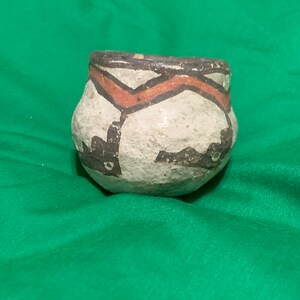 Könnte beinhalten: Kleines, rundes, handbemaltes Töpfergefäß mit einer weiten Öffnung. Der cremefarbene Körper ist mit einem Zickzackmuster in Orange und Schwarz verziert. Der Rand ist dunkelbraun. Das Gefäß steht auf einer grünen Oberfläche.