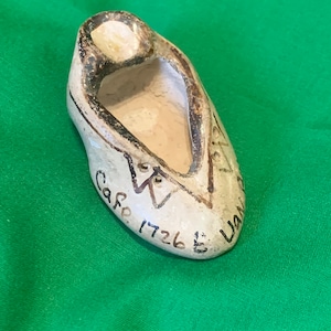 May include: A small, ceramic shoe figurine in off-white with brown trim and detailing. The shoe has a rounded toe and a small opening at the top. The text "Cafe 1726" is written on the side.