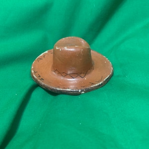 May include: A small, brown sombrero-shaped object with a cylindrical crown and a wide, flat brim. The brim has a decorative stitched pattern and some text. The hat is resting on a green surface.