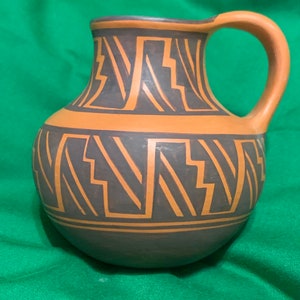 Op de afbeelding: Een handgemaakte aardewerken kruik met een afgeronde basis en een gebogen handvat. De kruik heeft een donkergrijze body met geometrische oranje ontwerpen en een bruin gekleurde rand en handvat. Het ontwerp doet denken aan Indiaanse kunst.