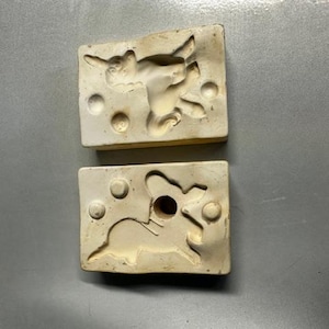 Puede incluir: Dos moldes rectangulares de cerámica blanquecina con diseño de conejo. Cada molde tiene una forma de conejo rebajada con tres hendiduras circulares. Los moldes probablemente se utilizan para fundir o crear objetos decorativos.