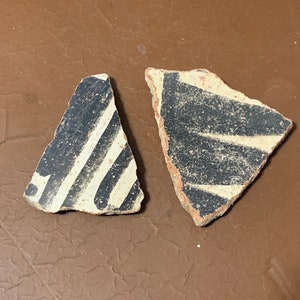 May include: Two fragments of ancient pottery with black and white striped designs. The pottery shards have a beige base with black and white stripes. The fragments are on a brown surface.