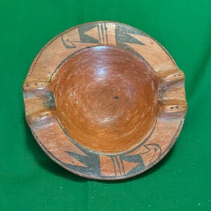 May include: A round, terracotta-colored ashtray with a central bowl and three cigarette rests. The rim features a black and tan geometric design. The ashtray is likely made of ceramic or clay.