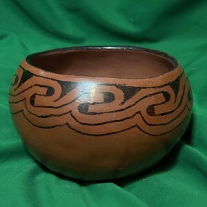 May include: A round, brown ceramic bowl with a dark interior. The exterior features a repeating black wave-like pattern around the rim. The bowl is set against a green background.