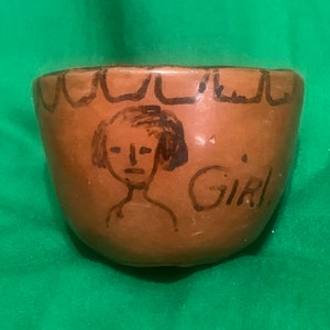 May include: A small, brown ceramic bowl with a hand-drawn illustration of a person and the word "GIRL". The bowl has a scalloped design around the rim. The illustration is in black ink.