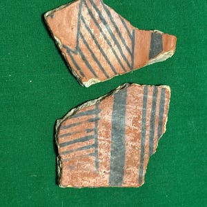 Puede incluir: Dos fragmentos de cerámica antigua con un color base marrón rojizo. Cada fragmento presenta un diseño de líneas negras diagonales y verticales. Los fragmentos de cerámica se muestran sobre un fondo verde.