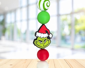 May include: A vertical beaded lanyard featuring a white bead with green swirls, a solid lime green bead, a central character bead depicting a green face wearing a red and white Santa hat, a solid red bead, and a solid white bead on a black cord.