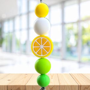Pode incluir: Uma corrente vertical de contas com uma esfera amarela brilhante, uma esfera branca, um pendente de fatia de citrino amarelo com um centro laranja texturado, uma esfera verde-lima e uma esfera verde-lima mais pequena em sequência.