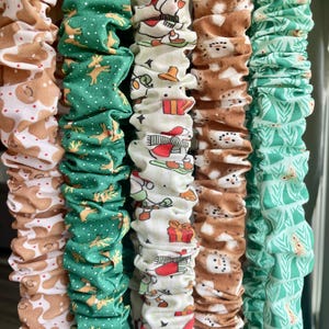 May include: Five fabric scrunchies arranged vertically. The first features brown gingerbread men on white, the second shows gold reindeer on green, the third displays holiday icons on cream, the fourth has brown snowmen, and the fifth is green.