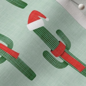 May include: A light mint green fabric featuring a repeating pattern of green cacti with vertical stripes. Each cactus is adorned with a red Santa hat and a red ribbon wrapped around its midsection. The fabric has a visible woven texture throughout.