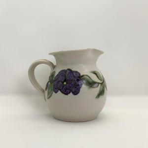 May include: A ceramic pitcher with a cream-colored base, featuring a purple floral design with green leaves. The pitcher has a handle and a spout for pouring. The floral design is hand-painted.