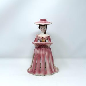 May include: A vintage ceramic figurine of a woman in a pink dress and hat, holding a tray. The figurine has a detailed face and is painted with pink, white, and brown colors. The dress has vertical cutouts.