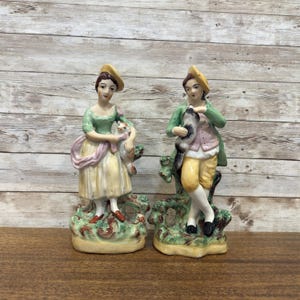 May include: A pair of vintage porcelain figurines depicting a man and woman in 18th-century clothing. The woman is holding a small white dog and the man is holding a bird. Both figurines are standing on a green base with brown accents.