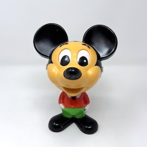 May include: A vintage Mickey Mouse figurine with a black head and ears, a tan face, and a red shirt. The figurine has green shorts and black shoes. The figure is standing on a white surface.