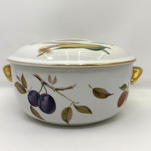 May include: White ceramic serving dish with lid, featuring a gold rim and handles. The dish is decorated with illustrations of plums, leaves, and corn. The lid has a corn graphic. The dish measures approximately 8 inches in diameter.
