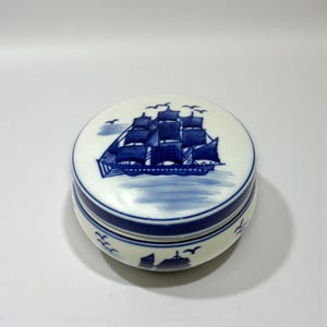 May include: A round, white ceramic trinket box with a blue nautical ship design on the lid. The box has a blue band around the middle and a matching blue design of birds around the base.