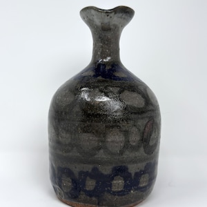 May include: A handcrafted ceramic vase with a dark gray base and a flared, gray-toned neck. The vase features a dark blue, patterned design around the base and upper body. The vase has a rustic, organic aesthetic.