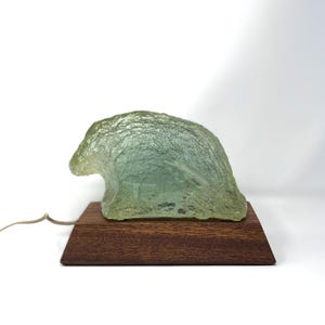 May include: A green glass lamp shaped like a wave, with a textured surface, sits on a wooden base.