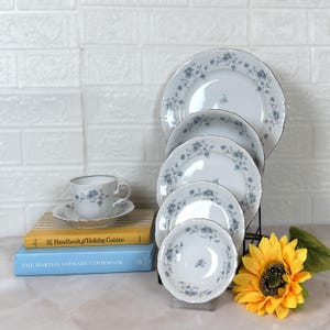 May include: A white porcelain dinnerware set with a blue floral pattern. The set includes plates, bowls, and a teacup with a saucer. The plates have a scalloped edge and a silver trim. A yellow sunflower is in the foreground.