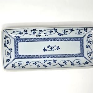 May include: Rectangular white ceramic serving dish with blue floral patterns. The dish has a rectangular shape with rounded corners. The border and interior are decorated with blue floral and vine motifs. The dish is ideal for serving appetizers or small dishes.