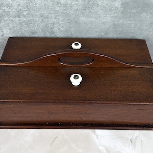 May include: A rectangular wooden box with a dark brown finish. It features a curved handle on top and two white ceramic knobs. The box is likely used for storage and has a classic, vintage aesthetic.