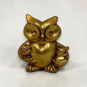 May include: A gold-colored owl figurine with two smaller owls on its sides. The owl has large eyes with yellow accents. The figurine is made of a textured material and sits on a green base. The owl is a decorative item.
