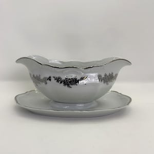 May include: White porcelain gravy boat with a matching saucer. The boat has a scalloped rim and is decorated with a floral pattern in silver and black. The edges are trimmed with silver.