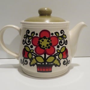 Puede incluir: Tetera de cerámica color crema con diseño floral. La tetera presenta una gran flor roja con centro amarillo, hojas verdes y volutas negras. La tapa y el asa son de color verde apagado.