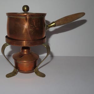Pode incluir: Uma panela de fondue de cobre com uma alça de madeira e um suporte de latão. A panela tem uma tampa com um pequeno botão de latão. O suporte tem pernas curvas e uma pequena base para o queimador. Um item de cozinha vintage.