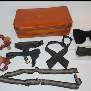 May include: A collection of men's accessories, including braces, bow ties, and neckties. A brown leather case is at the top, with black and striped items below. The bow ties are black, one in a clear case.