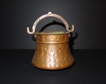 Puede incluir: Una pequeña olla de cobre martillada con asa. La olla tiene un fondo redondeado y un borde acampanado. El asa es de metal y tiene un diseño sencillo.