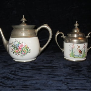 May include: A white porcelain teapot and sugar bowl set with silver-colored metal lids and handles. The teapot features a floral design, while the sugar bowl depicts a bird in a marsh setting. The set is likely vintage.