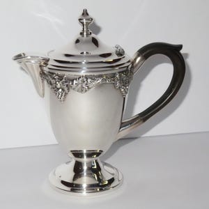 Puede incluir: Una cafetera de metal plateado con acabado pulido, tapa extraíble con remate y un asa curva de color marrón oscuro. La parte superior del cuerpo presenta una banda en relieve detallada con racimos de uvas y hojas sobre una base.