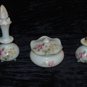 May include: A three-piece vanity set featuring a tall perfume bottle with a pointed stopper, a lidded powder jar, and a smaller perfume bottle with a gold cap. Each piece is made of white glass with hand-painted pink, yellow, and white rose floral motifs.