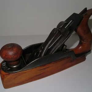 Peut inclure: Un rabot à main en bois avec une lame en métal et une poignée en bois. Le rabot est en bon état et a une finition brun foncé.