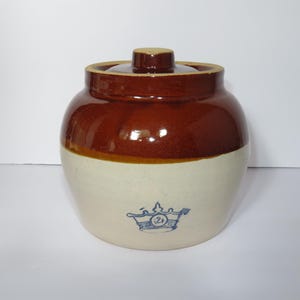 Op de afbeelding: Een vintage steengoed pot met een deksel. Het bovenste gedeelte is glanzend bruin, terwijl de onderste helft crèmekleurig is. Een blauw embleem is zichtbaar aan de voorkant. De pot is ontworpen voor opslag.