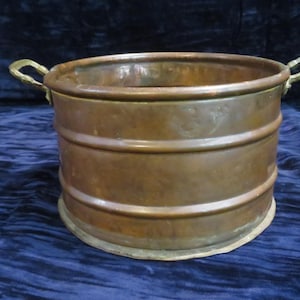 May include: A round copper basin featuring a smooth, metallic surface with a warm, reddish-brown patina. The vessel includes two small, curved brass handles attached to opposite sides and two horizontal raised bands encircling the exterior.
