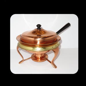 Könnte beinhalten: Ein Kupfer-Chafing-Dish mit Deckel und schwarzem Griff. Das Gericht hat ein messingfarbenes Band und steht auf drei gebogenen Beinen. Der Deckel hat einen schwarzen Knopf. Ein Klassiker zum Servieren von Speisen.