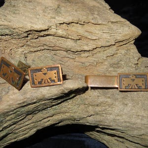 Pode incluir: Um conjunto de botões de punho e um prendedor de gravata com um design nativo americano estilizado. Os botões de punho e o prendedor de gravata são feitos de cobre e têm uma pátina marrom escura. O design é de um pássaro com as asas abertas.