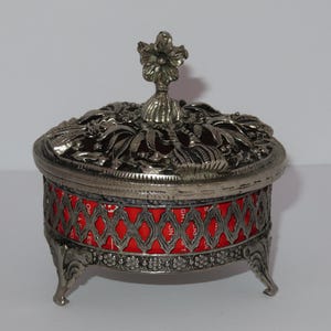 Può includere: Scatola portagioie argentata ornata con un motivo floreale sul coperchio e una fascia rossa e argentata. La scatola ha tre piedini decorativi e una maniglia a forma di fiore.