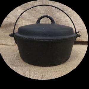 Peut inclure: Une cocotte en fonte noire avec un couvercle et une poignée. Le pot a une forme arrondie et une poignée sur le couvercle. La poignée est en fil métallique. Le pot est posé sur une surface en toile de jute.